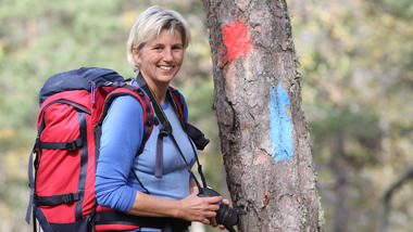 Smilende dame står ved et tre med sekk på ryggen og camera i hånden.