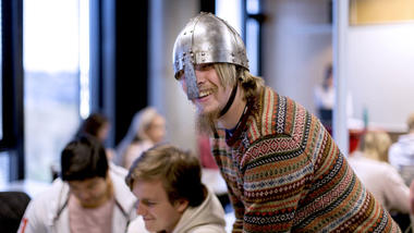 Mannlig lektorstudent i historie står i et klasserom i en undervisningssituasjon med en vikinghjelp hode og smiler til kamera. 