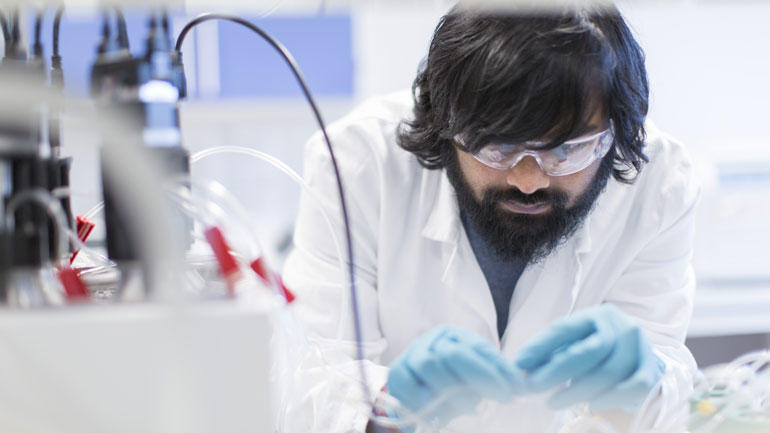 En mørkhåret student i hvit forskerfrakk og blå plasthansker jobber konsentrert i lab. 