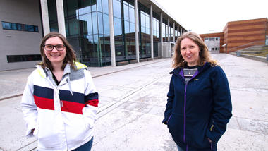 Hanna og Karina står utenfor i\hovedinngang til campus Vestfold og smiler til kamera. 