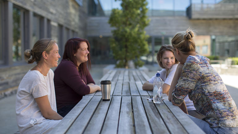 Fire studenter  sitter rundt et bord utendørs og snakker med hverandre og med termokoppen foran seg  i Kristinas Hage ved campus Vestfold.