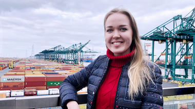 Mariia i rød høyhalsa genser og dunjakke, langt, lyst hår smiler med tenner til kamera foran et havnemiljø. 