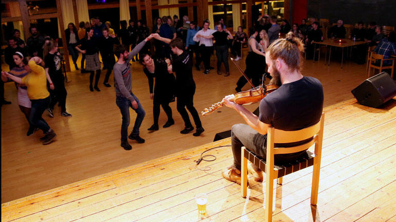 Ung mann speler fele på ei scene emdan folk dansar på golvet foran han. Foto