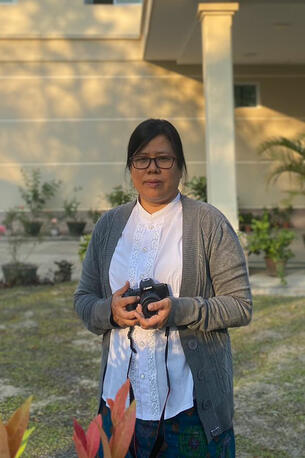  Picture of Thida Swe looking into camera, dark hair and glasses, holding a camera, white blouse and gray knitted jacket