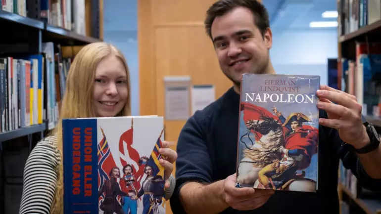 To studenter i bibliotek holder bøker og smiler mot kamera.