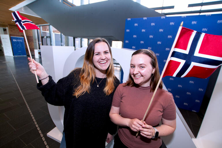 Lærerstudentene Linda g Aleksandra oppstilt innendørs ved USN-logi og med hvert sitt norske flagg i hånda, ser rett i kamera