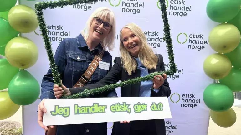 To smilende kvinner holder en ramme dekorert med grønt gress, og en plakat hvor det står "Jeg handler etisk, gjør du?" foran en bakgrunn med logoen til Etisk Handel Norge, omgitt av grønne ballonger.