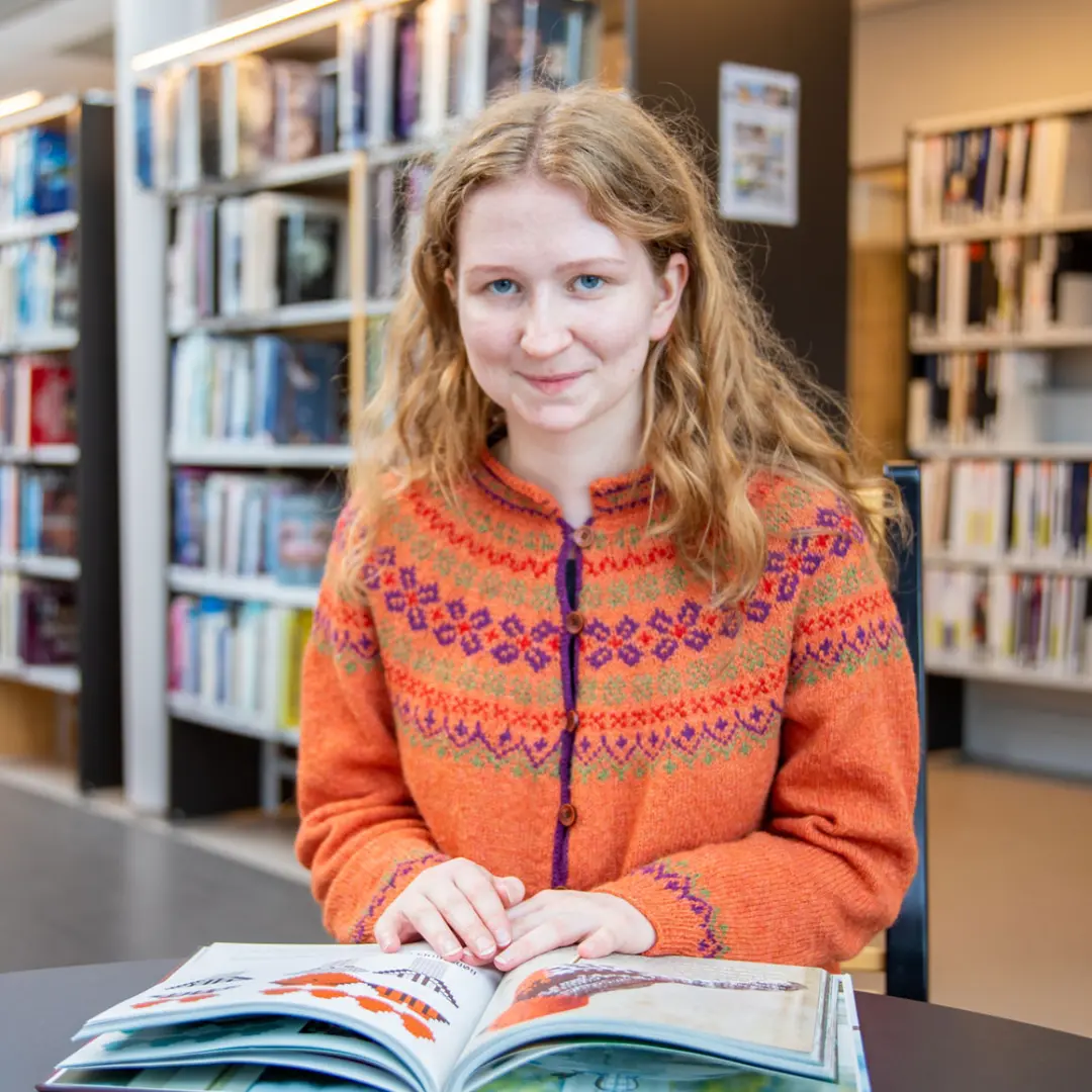 Ung kvinne i fargerik genser leser bok på biblioteket, omgitt av bokhyller fylt med forskjellige titler.