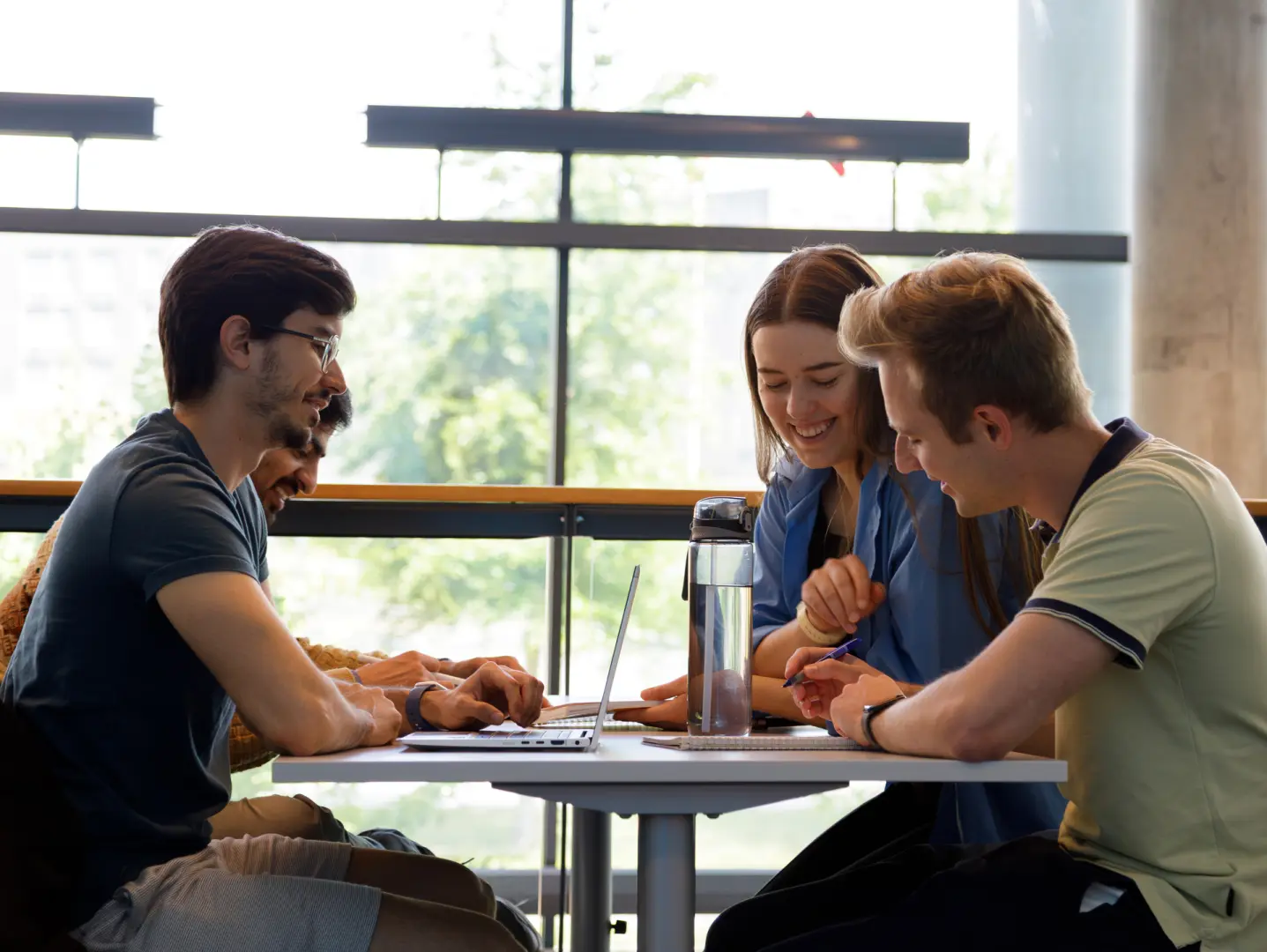 Gruppe studenter samarbeider ved et bord med bærbar PC og vannflaske, sittende ved store vinduer i et lyst innendørsrom.