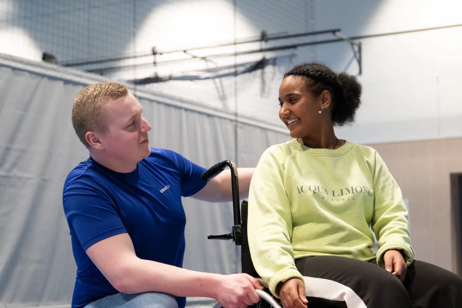 Vernepleierstudentene Halvard Tveitan og Yohana Foto Berhane i gymsal. Yohana sittende i rullestol.