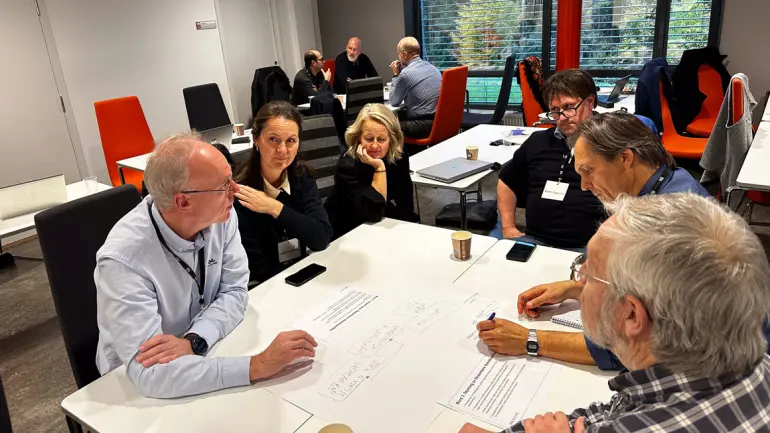 gruppe voksne mennesker diskuterer rundt et bord og noterer på ark