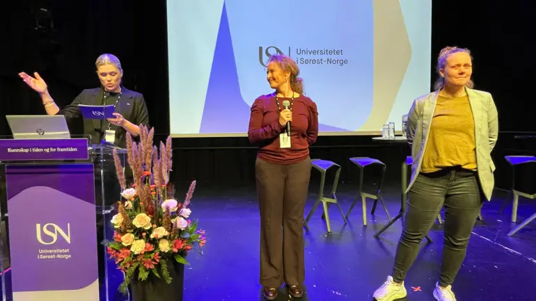 tre damer på en konferansescene pyntet med blomster og logoer for USN