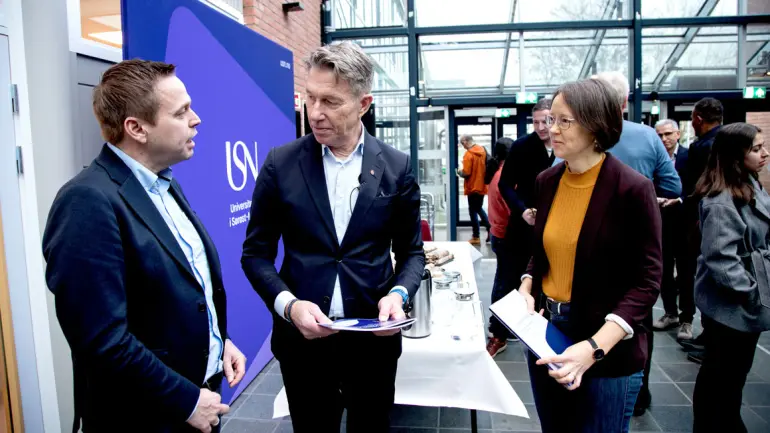 Halvfigur av energiminister Terje Aasland som snakker med forskerne Thomas Øyvang og Elin Fjeld ståenede i Glassgata med folk i bagrunnen på campus på Porsgrunn, og der Aasland holder rapportene i hendene. 