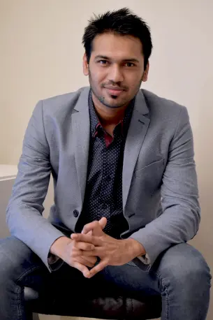 Portrett av Rocky Mahmud sittende i grå dress og med hendene på lårene og foldet  