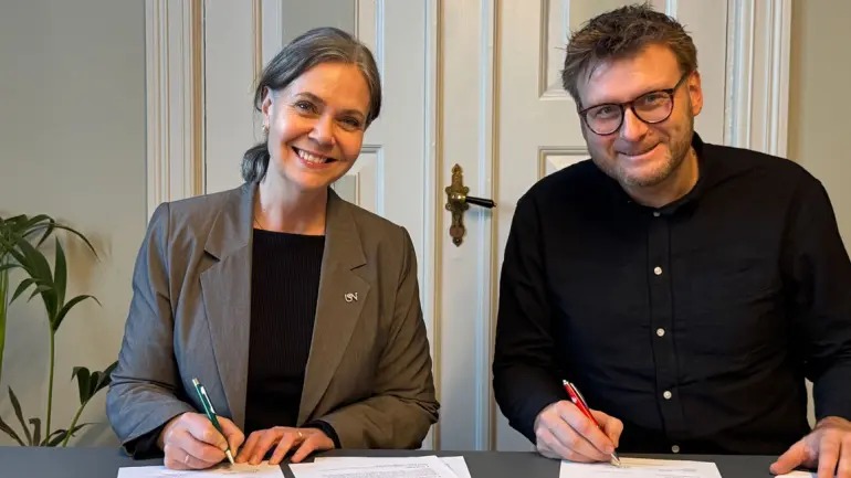 smilende mennesker bak bord med penn i hånden og papiret på bordet foran seg, mann og kvinne