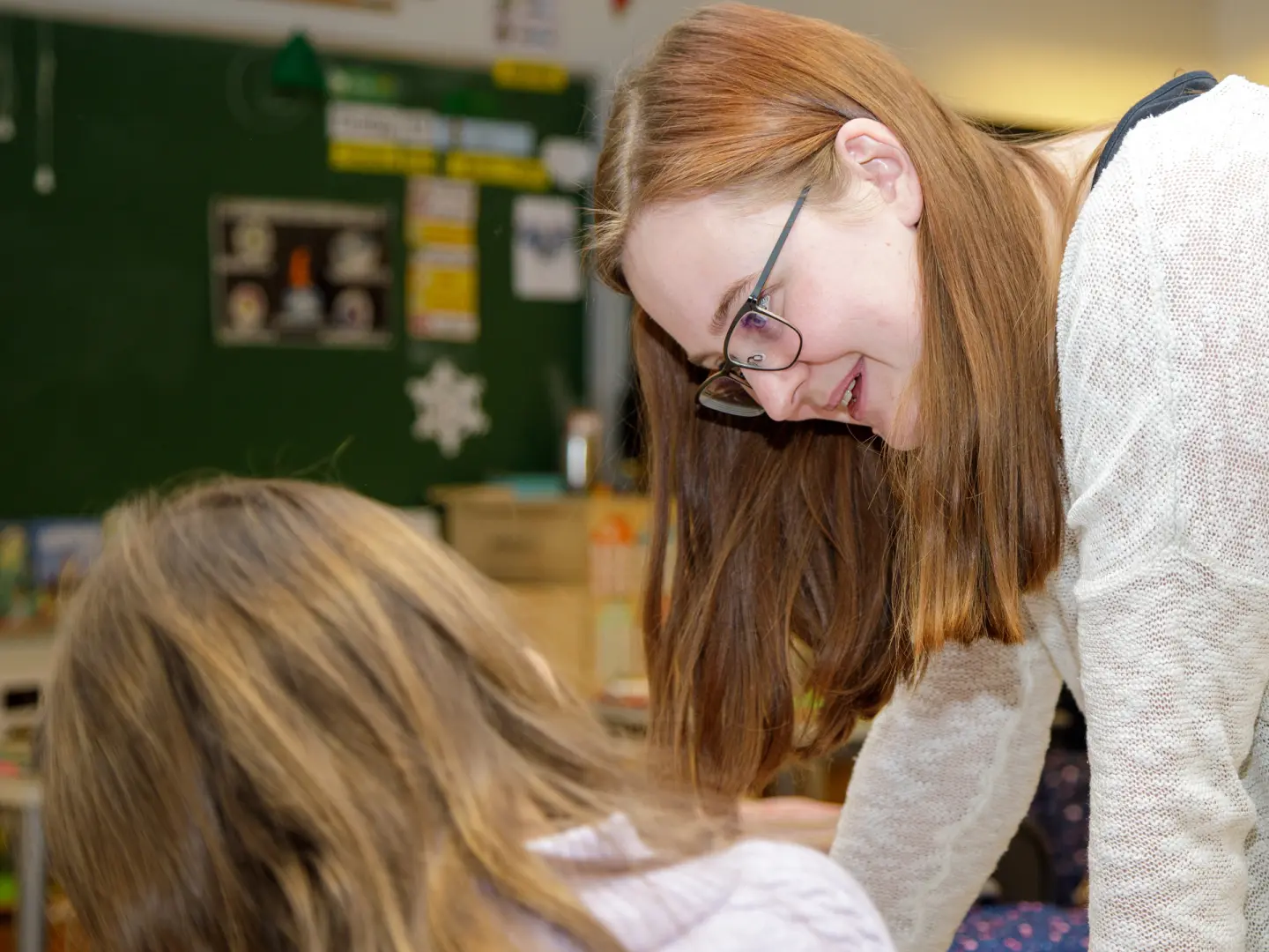  Lærer med briller og langt hår smiler mens hun hjelper en elev i et klasserom med grønn oppslagstavle.