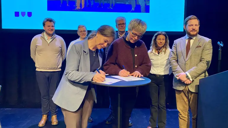 damer signerer papirer på et bord. flere mennesker står bak og smiler