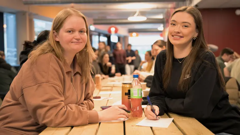 To studenter på seminar, lærerstudenter smiler inn i kamera mens de noterer.