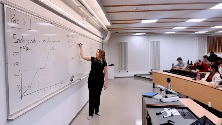 Karina i undervisningssituasjon foran tavla med studenter sittende og lytte i auditorium. 