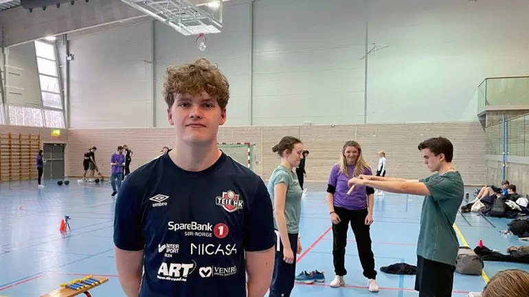 gutt i håndballdrakt ser i kamera i gymsal med aktivitet i bakgrunnen