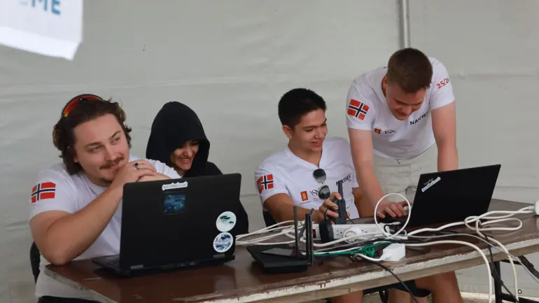 Studenter jobber ved et langbord med laptoper og styrer sjødronen på vannet herfra. 