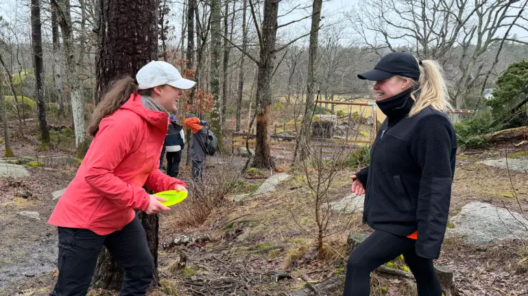 to damer ler sammen ute blant trærne, den ene holder en frisbee
