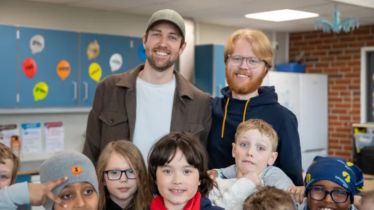Gruppebilde av to voksne og flere barn som smiler i et klasserom, med synlige fargerike skap og pedagogiske plakater.