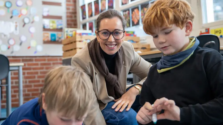 Kvinne med briller smiler med to unge gutter i et fargerikt klasserom med synlige kunstverk og undervisningsmateriell.