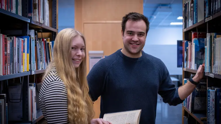 To smilende personer i en bibliotekgang, kvinne som holder en åpen bok, mann som berører en bokhylle. Omgitt av bokhyller.