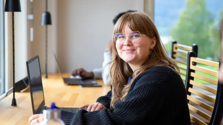 Kvinne med briller smiler til kameraet mens hun jobber på bærbar PC i et lyst kontorlokale med fjellutsikt.