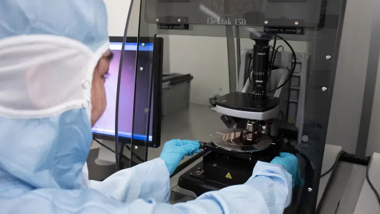 En forsker i beskyttelsesutstyr bruker et avansert mikroskop i laboratoriet og undersøker en prøve med et digitalt display i nærheten.