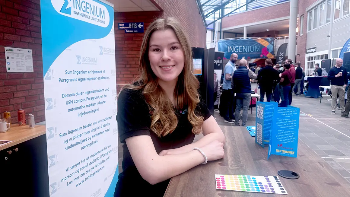 Mildrid ser i kamera mens bøyher seg over standen til linjeforeningen på karrieredagen for ingeniørstudenter på campus Porsgrunn 