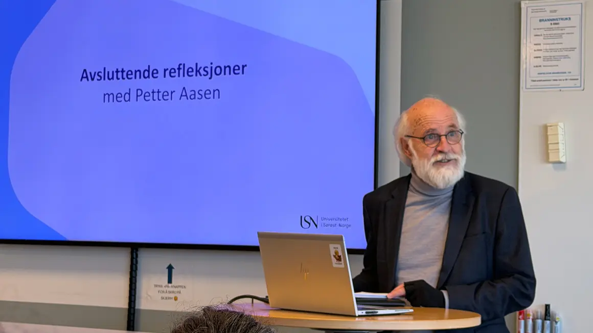 mann med skjegg presenterer foran tavle og skjerm