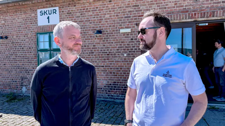 Halvportrett av Karl Thomoas Hjelmervik og TOrbjørn Hpuge som ser mot hverandre, stående i sola utendørs med murbygning i bakgrunnen.