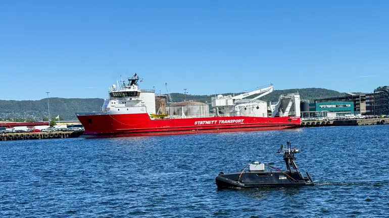 Et stort skip rødt og hvitt på havet og foran ligger det sorte autonome fartøyet som skal kunne avdekke mistenlig aktivtet