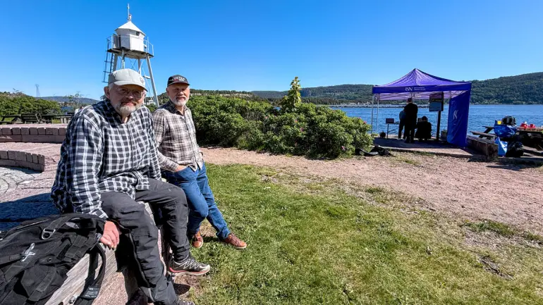 Helfigur av forskerne Jon Herman Ulvensøen og Geir Lislebø som sitter på en steinmur med USN-teltet og testområdet med havet i bakgrunnen 
