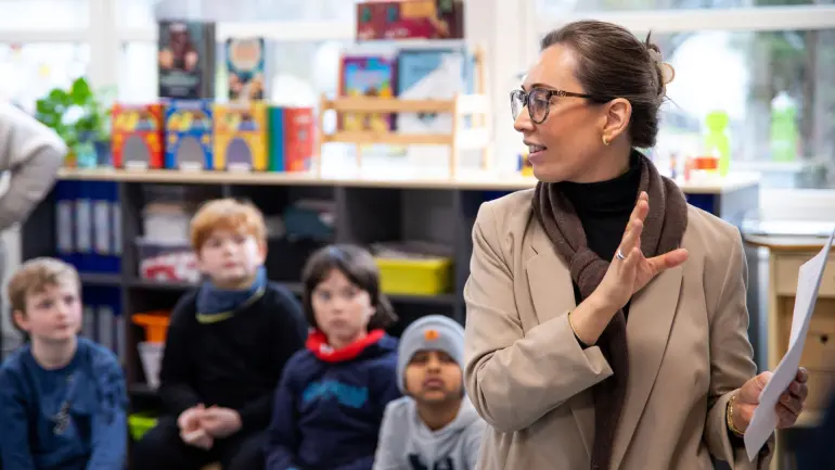Lærer gestikulerer mens hun snakker til oppmerksomme elever i et fargerikt klasserom med pedagogiske leker og materialer synlige. 