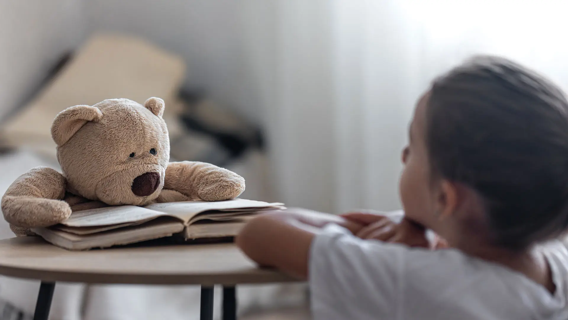 Barn med ryggen mot kamera ser på en bamse som sitter over en oppslått bok