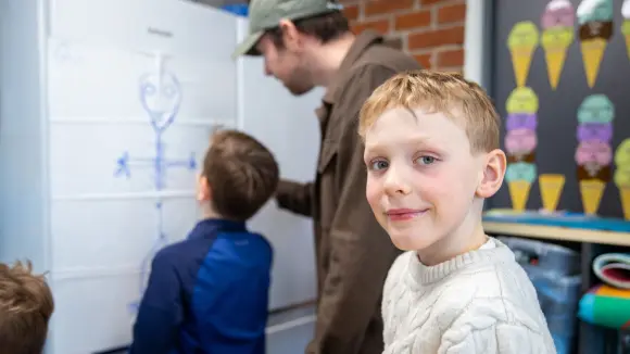 Barn i klasserom med lærer ved tavlen. Ung gutt i hvit genser smiler til kameraet. Fargerike iskrem-kunstverk er synlige.