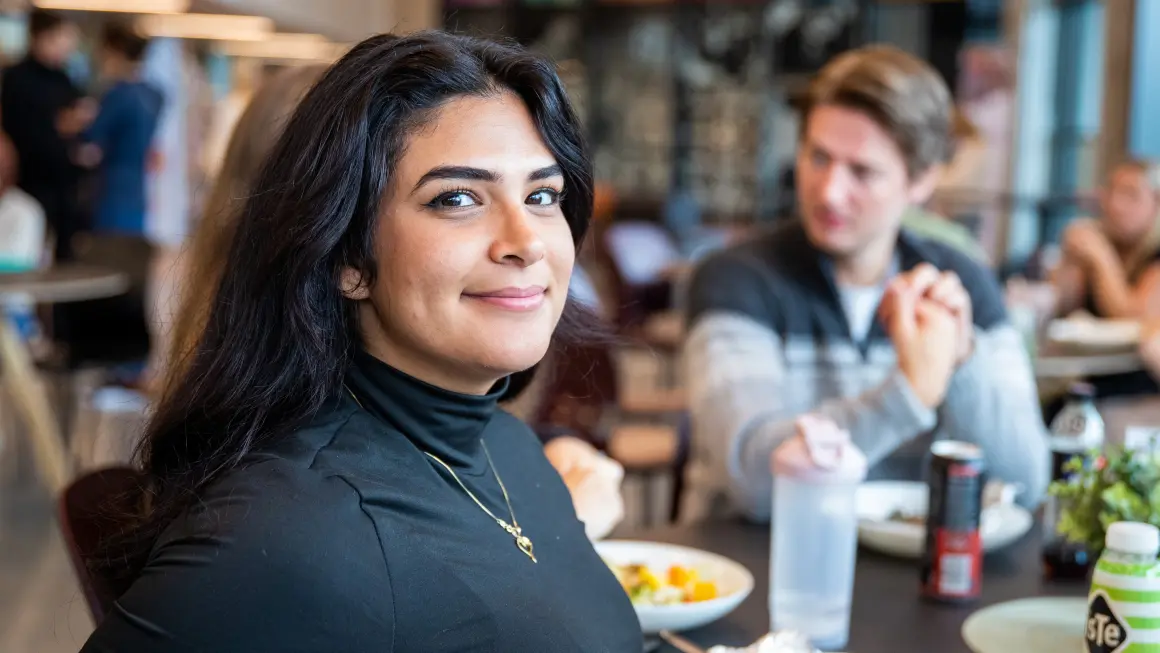 Student smiler til kamera i kantine, sammen med andre studenter som spiser lunsj i bakgrunnen.