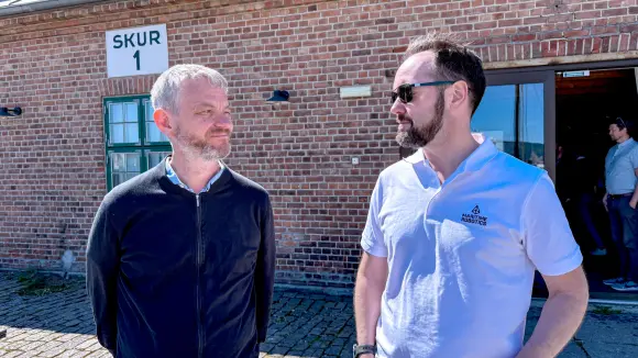 Halvportrett av Karl Thomoas Hjelmervik og TOrbjørn Hpuge som ser mot hverandre, stående i sola utendørs med murbygning i bakgrunnen.
