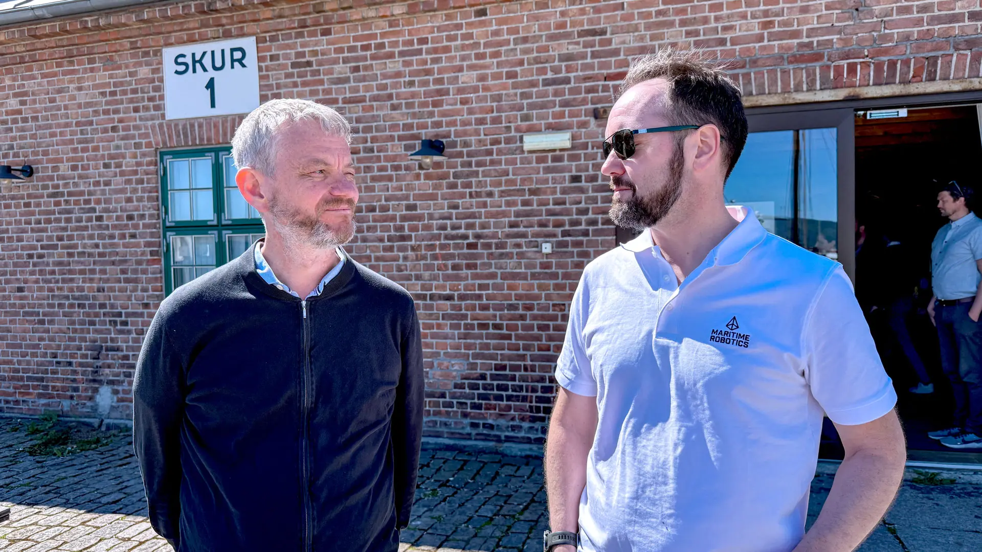 Halvportrett av Karl Thomoas Hjelmervik og TOrbjørn Hpuge som ser mot hverandre, stående i sola utendørs med murbygning i bakgrunnen.
