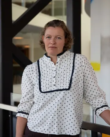 Gunhild Hustad i hvit mønstret bluse som står innendørs nær en døråpning.