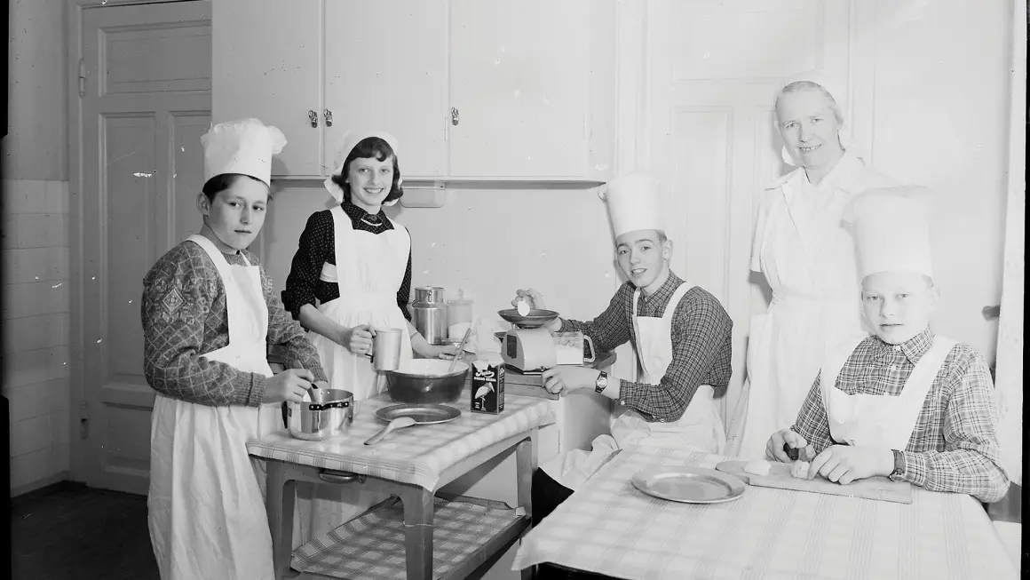 “Lykkelige og nyttige samfunnsborgere” var skolens motto. Elevene måtte framstå som veltilpassede og lykkelige når potensielle givere kom på besøk. Her fra skolens kjøkken på 1950-tallet. Gjengitt med tillatelse fra Vestfoldmuseene. Foto: Nøtterø foto En gruppe mennesker i forklær som lager mat på kjøkkenet