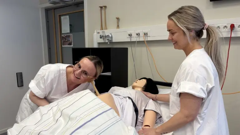 To studentar står ved sjukesenga og lytter til magen hos gravid kvinne.