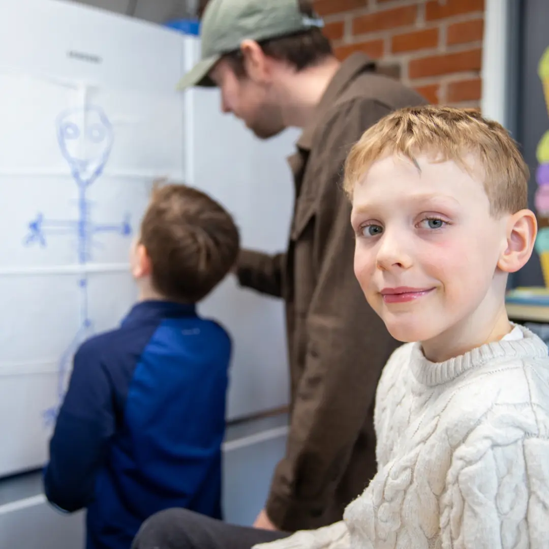 Barn i klasserom med lærer ved tavlen. Ung gutt i hvit genser smiler til kameraet. 