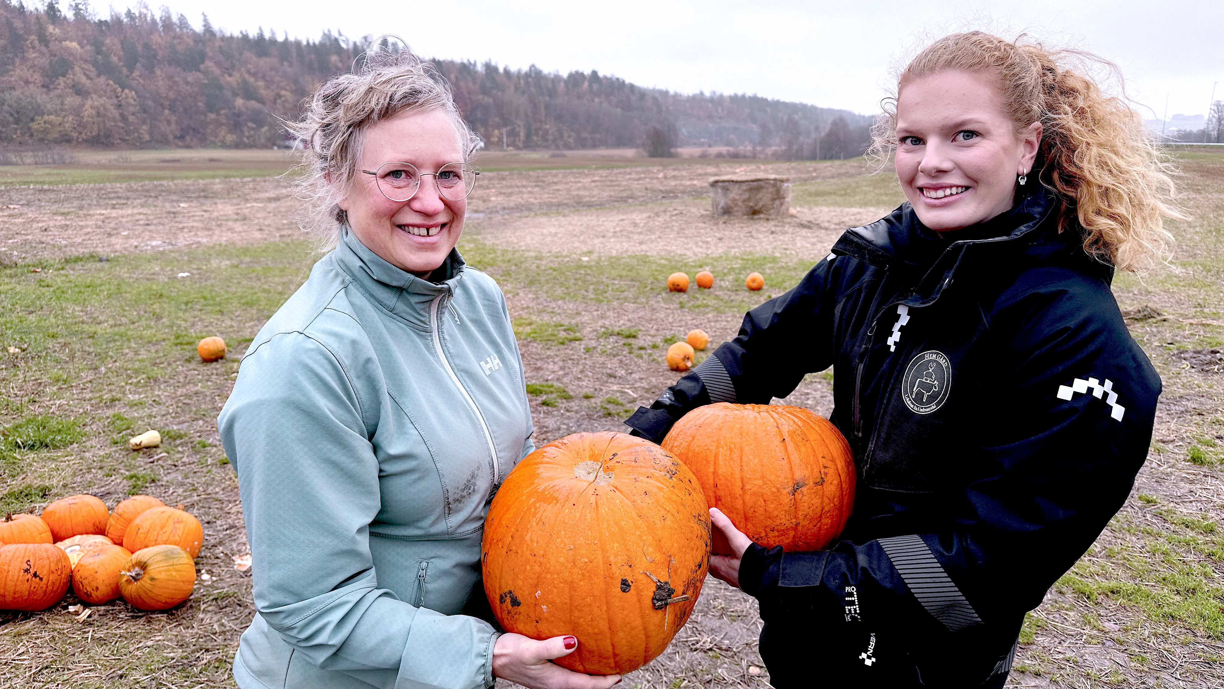 Portrett av USN-forsker Susanne Moen Ouff og odelsjente Solveig Eline Eikeberg stående ute på jordet på Hem Gård med hvert sitt oransje gresskar i hendene. 