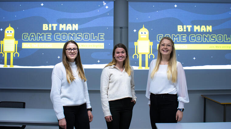 tre kvinnelige ingeniørstudnter står på en rekke og smiler til kamera med storskjerm i bakgrunnen 