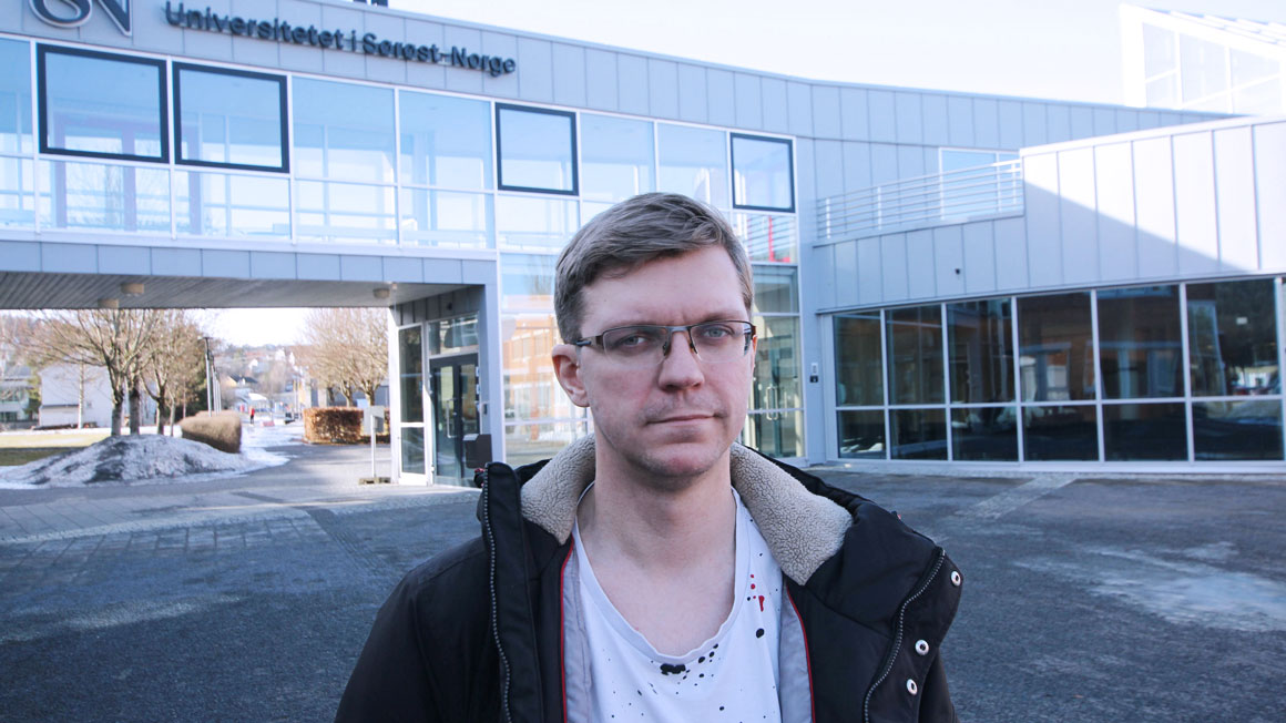 Portrett av Petter med kort, møkrt hår og briller stående ute med USN-bygningen i bakgrunnenn