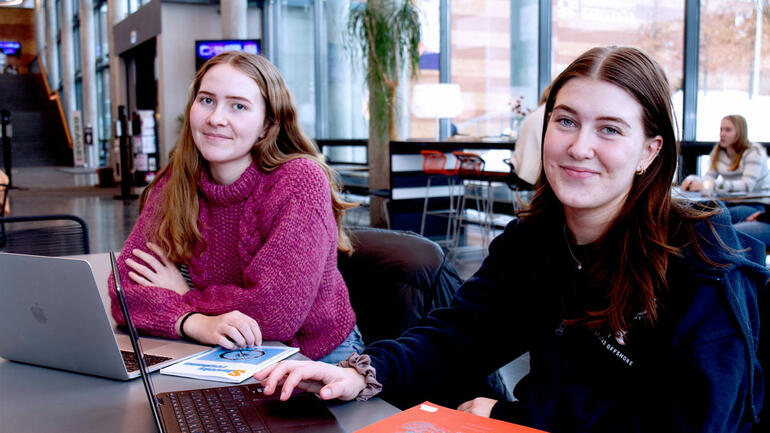 Teknologistudentene Jona og Julie sittende ved et bord med laptop og smiler til kamera